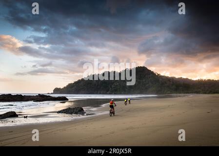 Ciclisti che cavalcano lungo una spiaggia deserta verso il tramonto, Giava, Indonesia Foto Stock