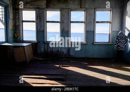 Un ragazzo nell'angolo di una stanza di una casa in legno abbandonata con finestre vuote senza vetro e vista sul fiume Foto Stock