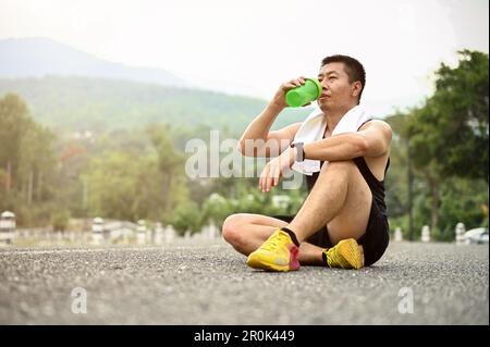 Uomo asiatico sudato e assetato in abbigliamento sportivo si siede sulla strada in un parco pubblico e beve acqua da una bottiglia dopo la corsa. Foto Stock