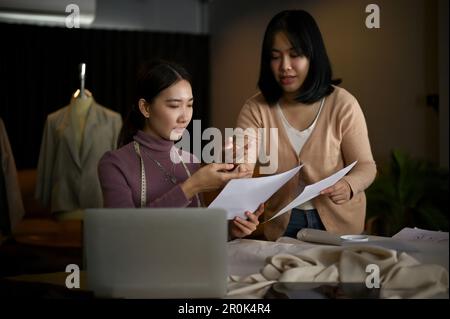 Due giovani stilisti o sarte asiatiche professionisti stanno discutendo la loro nuova collezione e lavorando insieme in studio. Foto Stock