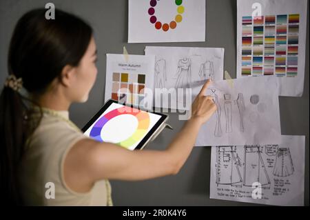 Designer di moda femminile asiatica professionale e creativa con il suo tablet digitale che controlla la nuova collezione di schizzi design nel suo studio. vista posteriore Foto Stock