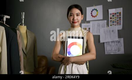 Ritratto di una fidata stilista asiatica di moda millennial che tiene il suo tablet con la carta a colori sullo schermo, in piedi nel suo studio creativo. Foto Stock