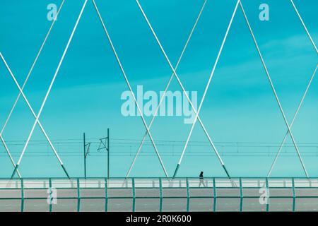 Una persona irriconoscibile che indossa una giacca nera con cappuccio sta attraversando una grande struttura a ponte durante il giorno di primavera soleggiato, fuoco selettivo Foto Stock