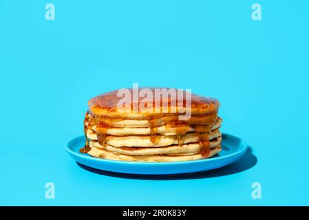 Pila di frittelle ricoperte di sciroppo su un piatto blu, minimalista su un tavolo blu. Deliziosi pancake fatti in casa con sciroppo d'acero in luce brillante su un co Foto Stock