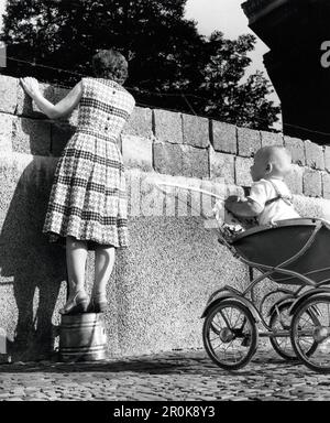 Eine junge Mutter, dahinter ihr kind in seinem Kinderwagen, schaut über die Mauer in der Bernauer Strasse in den Ostsektor von Berlin, Deutschland 1961. Foto Stock