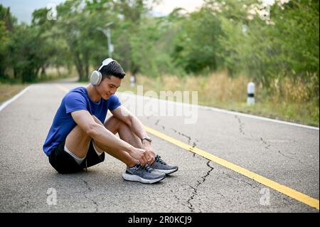 Felice uomo asiatico millenario che indossa le cuffie si siede sulla strada e lega i suoi lacci scarpe sportive, preparandosi per correre o fare jogging in un parco. Foto Stock