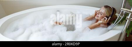 Caucasica giovane bella donna in cuffie sdraiato in vasca da bagno con schiuma e ascoltare musica Foto Stock