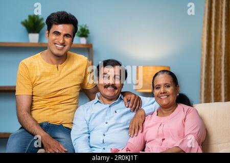 Ritratto di felice sorridente coppia indiana anziana con figlio adulto che guarda la macchina fotografica mentre si siede sul divano a casa - concetto di legame familiare Foto Stock