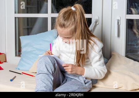 il bambino disegna su carta con penne in feltro, seduto sul davanzale nella stanza. La ragazza scrive un testo su carta. Il bambino sta facendo un lesso della scuola Foto Stock