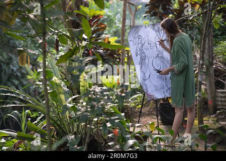 una donna dipinge un'immagine di un paesaggio, una ragazza è un artista, una vista posteriore, un parco estivo, un lago, uno stagno, un fiume che crea un'atmosfera artistica creativa. Foto Stock