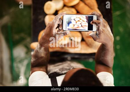 Caricare foto del mio pane è ciò che mi rende così popolare. un panettiere maschio che fotografa sul suo cellulare una selezione di pane appena sfornato. Foto Stock