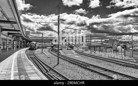 Tre treni aspettano in una stazione ferroviaria. Una passerella collega le piattaforme e un cielo con le nuvole è sopra. Un parcheggio auto è su un lato. Foto Stock