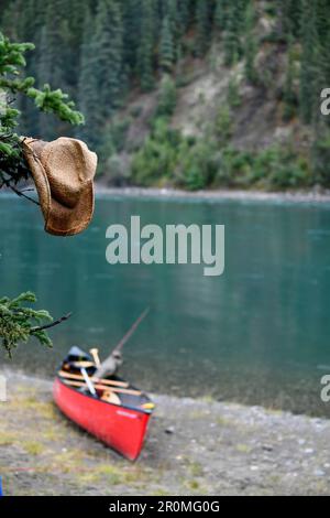 Cappello di paglia bagnato appende da un albero, sullo sfondo una canoa sulla riva, pausa al fiume Yukon, Yukon, Canada Foto Stock