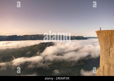 L'uomo sorge all'alba sul bordo di Preikestolen. Sotto di lui nuvole e il Lysefjord. Norvegia Foto Stock