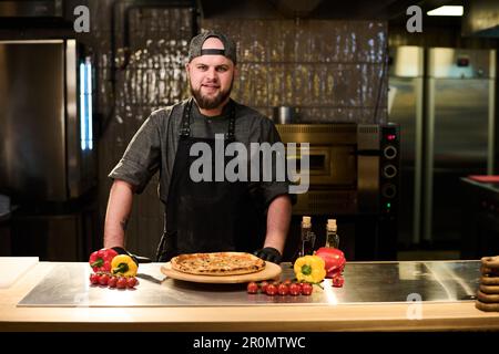 Happy young successful chef and owner of restaurant or pizzeria looking at camera while standing by table with appetizing pizza in the kitchen Foto Stock