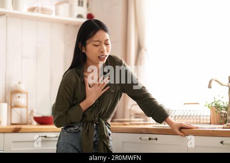 Donna asiatica malata che soffre di bruciore di stomaco in cucina, toccando il petto Foto Stock