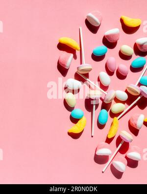 Vista dall'alto di varie gelatine e gommose colorate con caramelle smaltate e dure con lecca-lecca su bastoncini appoggiati su sfondo rosa Foto Stock