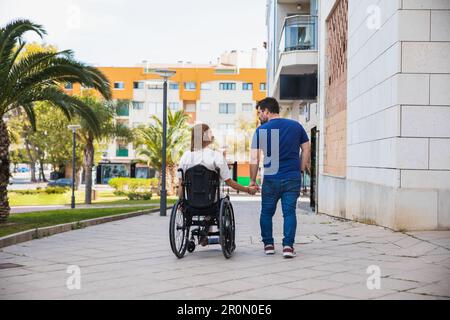 Vista posteriore del corpo pieno di marito allegro con moglie disabile in sedia a rotelle in abiti casual su strada piastrellata proveniente dalla clinica tenendo le mani e loo Foto Stock