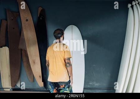Vista posteriore di anonimo padrone maschio tatuato in abiti casual in piedi in officina moderna e guardando la tavola da surf mentre si controlla la qualità del lavoro Foto Stock