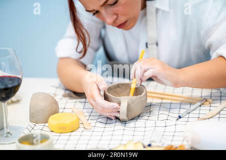 Ritaglia giovane artigiana focalizzata in grembiule utilizzando un bastone speciale per levigare la tazza in ceramica mentre crei terracotta fatta a mano in studio professionale Foto Stock