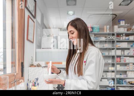 Giovane giovane farmacista in vestito bianco utilizzando mortaio e pestello mentre si prepara ingrediente medico durante la giornata di lavoro in farmacia Foto Stock