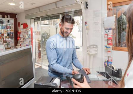 Crop femmina farmacista utilizzando scanner di codici a barre mentre servendo felice cliente maschile con codice QR sul telefono cellulare durante il pagamento contactless Foto Stock