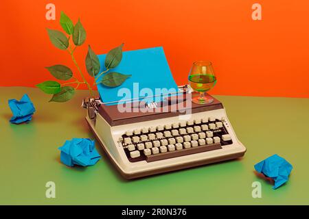 Alto angolo di macchina da scrivere d'epoca con foglie verdi e carte blu stropicciate con bicchiere di vino su sfondo verde e arancione in studio Foto Stock