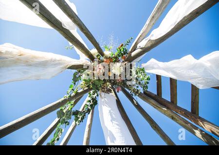 Decorazione rustica per matrimoni, arco da cerimonia con fiori bohemien, tessuto bianco e legno su sfondo cielo blu. composizione floreale su scala in legno Foto Stock