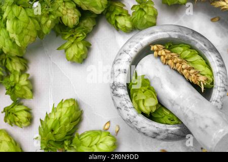 Mortaio con pestello, luppolo fresco e spighe di grano su tavola in marmo grigio chiaro, piatto Foto Stock