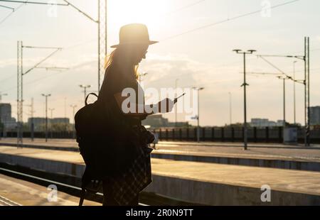 Donna turistica con zaino in piedi sulla piattaforma della stazione ferroviaria e tenendo il telefono in mano contro il tramonto. Foto Stock