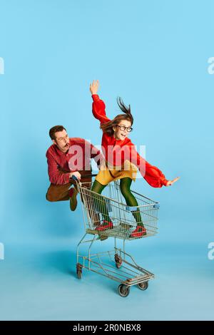 Ritratto con coppia divertente, bell'uomo e donna eccitata a cavallo nel carrello della spesa su sfondo blu studio. Vendite del Black Friday Foto Stock