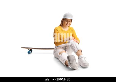 Giovane donna con uno skateboard seduto sul pavimento e tenendo il ginocchio ferito isolato su sfondo bianco Foto Stock