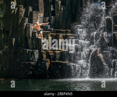 Ragazzo adolescente che salta in cascata lago. I corsi d'acqua che cadono scorrono sulle cascate di pietra vulcanica nera. Cascata di Rochester Falls - popolare punto turistico i Foto Stock