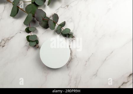 Vista dall'alto delle foglie di eucalipto e del podio bianco rotondo su sfondo marmoreo. Forma geometrica presentazione del prodotto cosmetico. Giacitura piatta, spazio di copia. Foto Stock