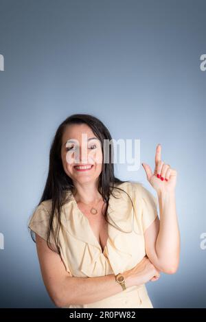 Una giovane donna adulta con lunghi capelli scuri, che indossa una t-shirt gialla casual, che punta con il dito indice mentre si trova su uno sfondo bianco Foto Stock