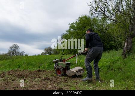 Un uomo aziona un motocoltivatore sul campo, coltivando il terreno arato e preparandolo per la semina in un giorno di primavera. Concetto agricolo Foto Stock