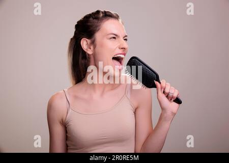 Bella donna che canta ad alta voce in spazzola durante la routine di acconciatura. Attraente giovane modello caucasico posato con pettine, utilizzando l'accessorio per capelli come microfono e divertendosi Foto Stock