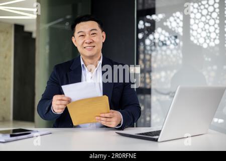 Ritratto di un uomo d'affari di successo all'interno di un moderno edificio di uffici, l'uomo asiatico ha ricevuto buone notizie in busta apre lettera felice sorridendo e guardando la fotocamera. Foto Stock