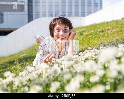 Donna in occhiali da sole colorati sniffs fiori trifoglio sul prato in parco urbano. Natura in città. Rilassati all'aperto dopo il lavoro. Atmosfera estiva. Foto Stock