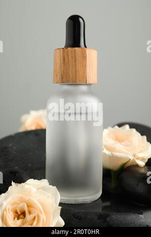 Bottiglia di siero facciale con belle rose su pietre spa su sfondo grigio, primo piano Foto Stock