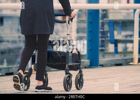 Donna anziana disabile cammina con il camminatore. Una donna che tiene un camminatore mentre cammina per la strada. Messa a fuoco selettiva, copyspace, foto di strada Foto Stock