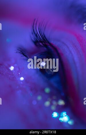 Vista ravvicinata estrema dell'occhio femminile con un make-up brillante con scintille e frecce. Bellezza notte macro ripresa di occhio femminile illuminato di colore rosa e blu Foto Stock