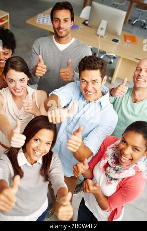 Appassionato e positivo per il loro servizio. Ritratto con vista ad angolo alto di un gruppo di persone multietniche che tengono i pollici in un ambiente di lavoro. Foto Stock
