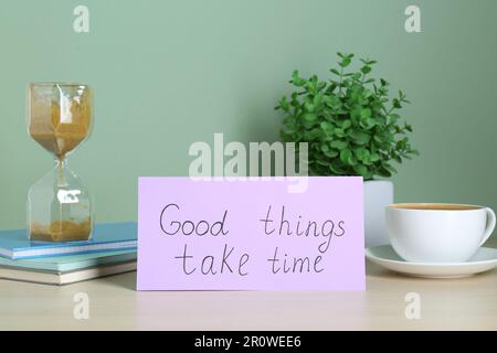 Frase buone cose prendere tempo, caffè, pianta della casa e clessidra sul tavolo contro sfondo verde chiaro. Citazione motivazionale Foto Stock