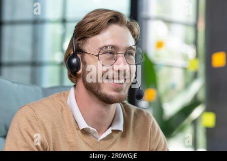 Uomo d'affari di successo con cuffie per videochiamate che lavorano all'interno dell'ufficio, un uomo che si occupa di consulenza online con i clienti, che parla con i colleghi durante una riunione remota online, che si siede sul posto di lavoro con un computer portatile Foto Stock