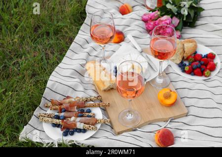 Bicchieri di delizioso vino di rosa, fiori e cibo su coperta da picnic all'aperto Foto Stock