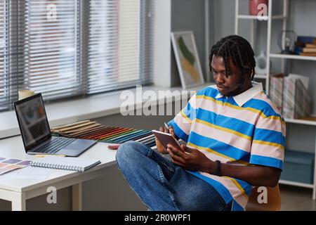 Giovane designer afroamericano che disegna un progetto su un tablet pc seduto sul suo posto di lavoro in ufficio Foto Stock