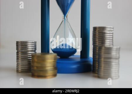 La sabbia blu che cade in un vetro di sabbia circondato da colonne di monete illustra il prezzo del tempo Foto Stock