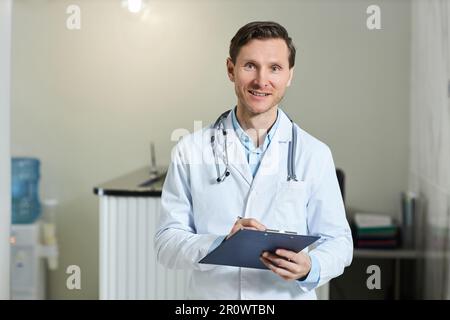 Vita in su ritratto di uomo sorridente come medico che indossa camice da laboratorio in clinica e guardando la fotocamera tenendo appunti, spazio copia Foto Stock