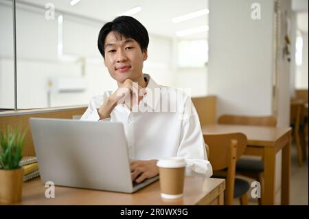 Giovane asiatico elegante e attraente in camicia bianca siede in una caffetteria con il suo computer portatile e la sua tazza da caffè su un tavolo. Foto Stock